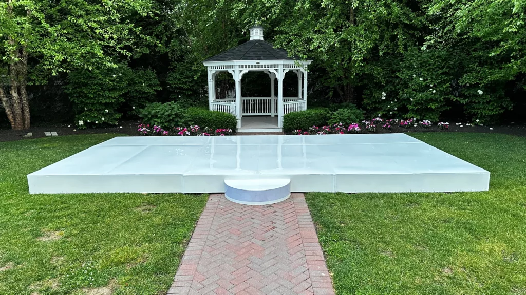 A white gazebo located at the end of a brick pathway, surrounded by neat green shrubbery and pink flowers, with a large, flat white stage in the foreground.