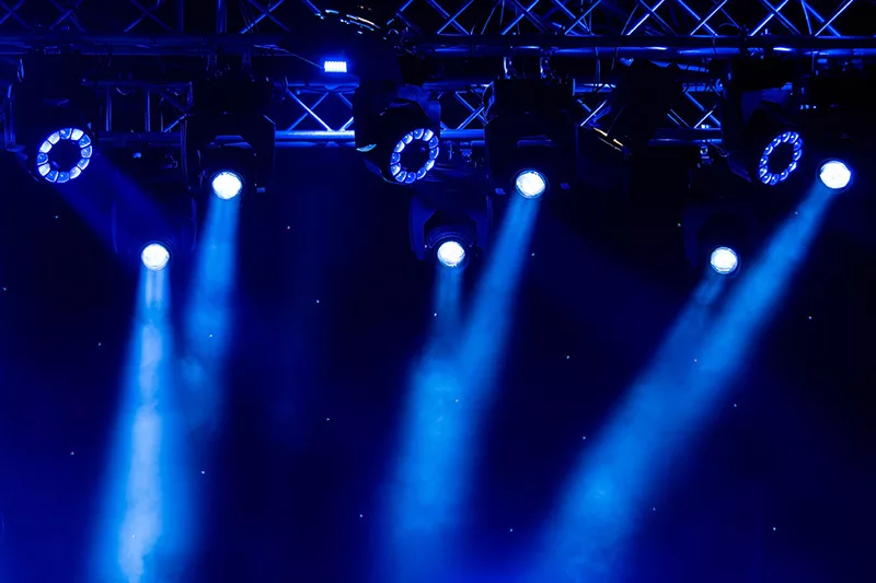 Stage lights with blue beams shining downwards from a truss at a concert or event, against a dark background.