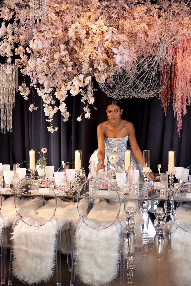 Model in shiny blue dress standing in front of Elegant Wedding Furniture Rentals such as a clear table with clear chairs and fur seats with a display of floral arrangement on the table and draping from the top of the ceiling