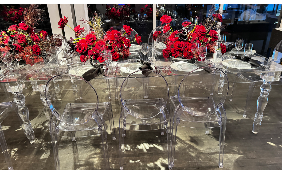 Elegant dining table setup featuring clear acrylic chairs and multiple bouquets of red flowers, with detailed table settings including plates, glasses, and cutlery.