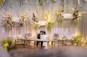A couple dressed in formal attire shares a kiss at an elegantly decorated wedding table. The table is adorned with numerous candles and floral arrangements in white, pink, and green hues. The background features hanging floral decorations, creating a romantic atmosphere.