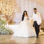 A bride in a white gown and a groom in a white tuxedo walk hand in hand at a wedding. They are surrounded by elaborate floral arrangements in white and gold. The setting is elegant, with soft lighting and a draped backdrop.