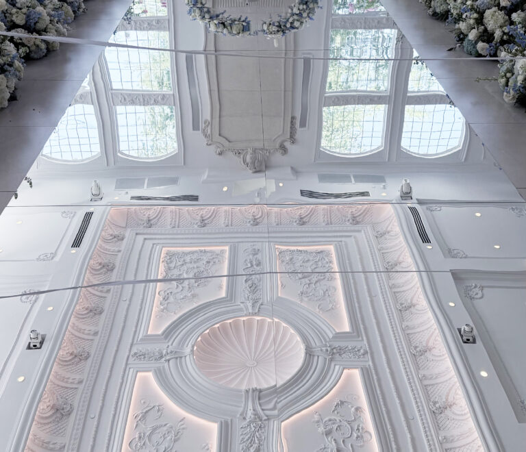 Elegant mirrored wedding aisle with floral arrangements. A wedding altar decorated with abundant white flowers and candles reflects beautifully on a glossy acrylic white aisle, while white chairs and draped gray curtains complete the elegant indoor setting.