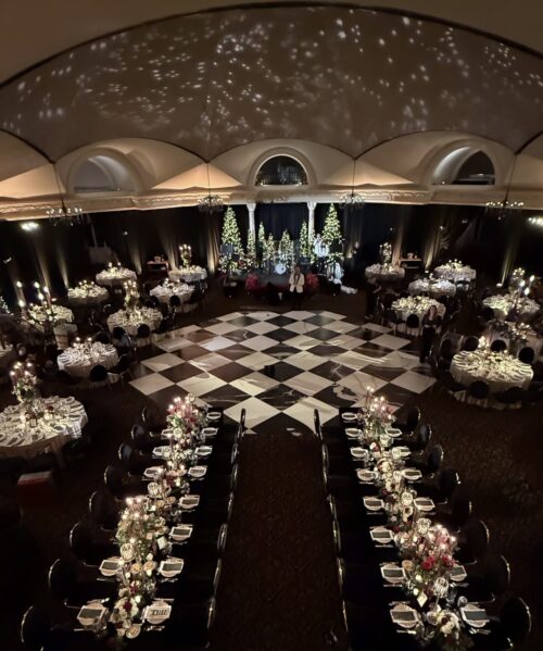 Elegant banquet hall with round and long tables decorated with flowers and candles, a black-and-white checkered dance floor, arched ceiling, and Christmas trees with lights at the back; soft, warm lighting throughout.