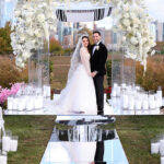 A bride and groom stand under a white floral wedding arch, surrounded by candles, on a mirrored aisle outdoors with a city skyline in the background.