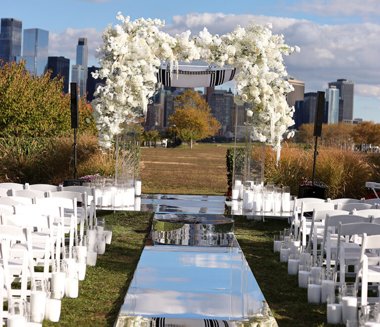 Luxury mirror aisle rental at New Jersey wedding venue. An outdoor wedding aisle lined with white chairs and candles leads to a floral arch with white flowers, mirrored walkway, and a city skyline visible in the background under a blue sky with clouds.