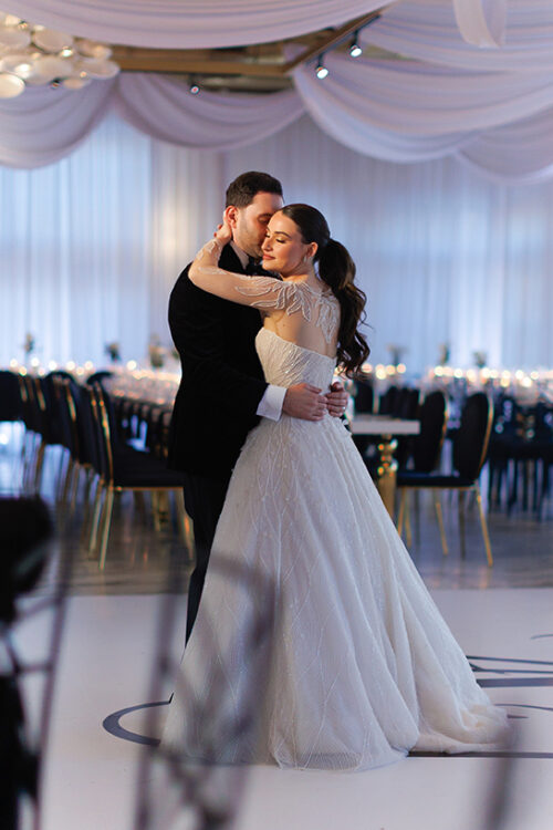 A bride in a white gown and a groom in a dark suit share a close, intimate dance on a decorated ballroom floor, surrounded by empty chairs and tables set for an event.