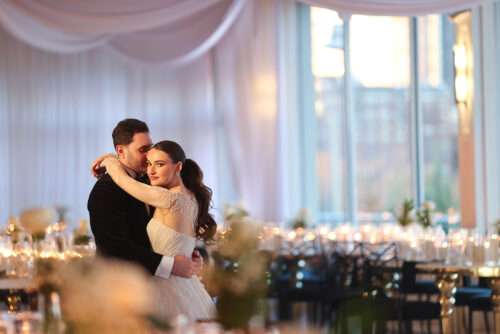 A bride and groom embrace on the dance floor in a softly lit, elegant wedding venue with draped white fabric, large windows, and tables decorated with candles and flowers.
