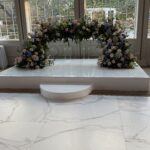 A white platform with a small round step features a lush floral arch made of pastel flowers in front of large windows, with trees visible outside. The setting suggests a wedding or formal event.
