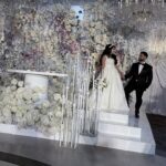 A bride and groom hold hands, smiling as they step down white stairs decorated with white flowers and tall candles. Elegant white drapery and a large floral arrangement create a luxurious wedding backdrop.