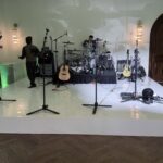 A stage setup with microphones, guitars, and a drum set. A person stands to the left while a drummer is seated. Green lighting and wood floors create a warm atmosphere, with a projection screen visible on the far left.