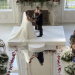 A bride and groom stand on a white platform exchanging vows in a bright, elegant room decorated with flowers and rose petals, with their reflection visible on a shiny floor. Guests are seated on either side of the aisle.