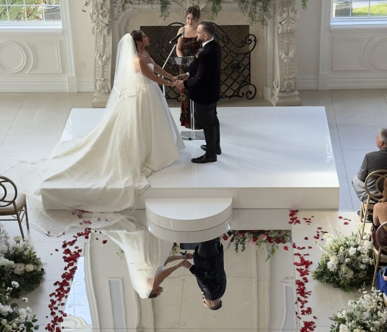 Custom mirror aisle installation by Class Event Rentals. A bride and groom stand on a white platform exchanging vows in a bright, elegant room decorated with flowers and rose petals, with their reflection visible on a shiny floor. Guests are seated on either side of the aisle.