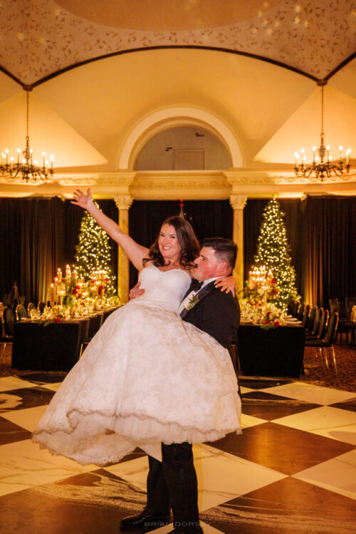 A groom in a suit lifts his bride in a white wedding dress on a black-and-white checkered dance floor, both smiling joyfully in an elegant hall with chandeliers and decorated Christmas trees.