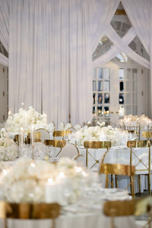 A luxurious event setup with round tables covered in white linens, gold chairs, white floral centerpieces, candles, and elegant glassware. White drapes and large windows create a bright, sophisticated atmosphere.