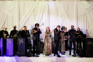 A band of nine performers in formal, glittering attire sings and plays instruments on stage in front of white draped curtains, with microphones and music stands arranged across the stage.
