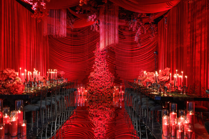 A lavish event space decorated in deep red drapes, with a mirrored floor, long tables set with candles and flowers, and a large floral arrangement at the center, creating a dramatic, romantic atmosphere.