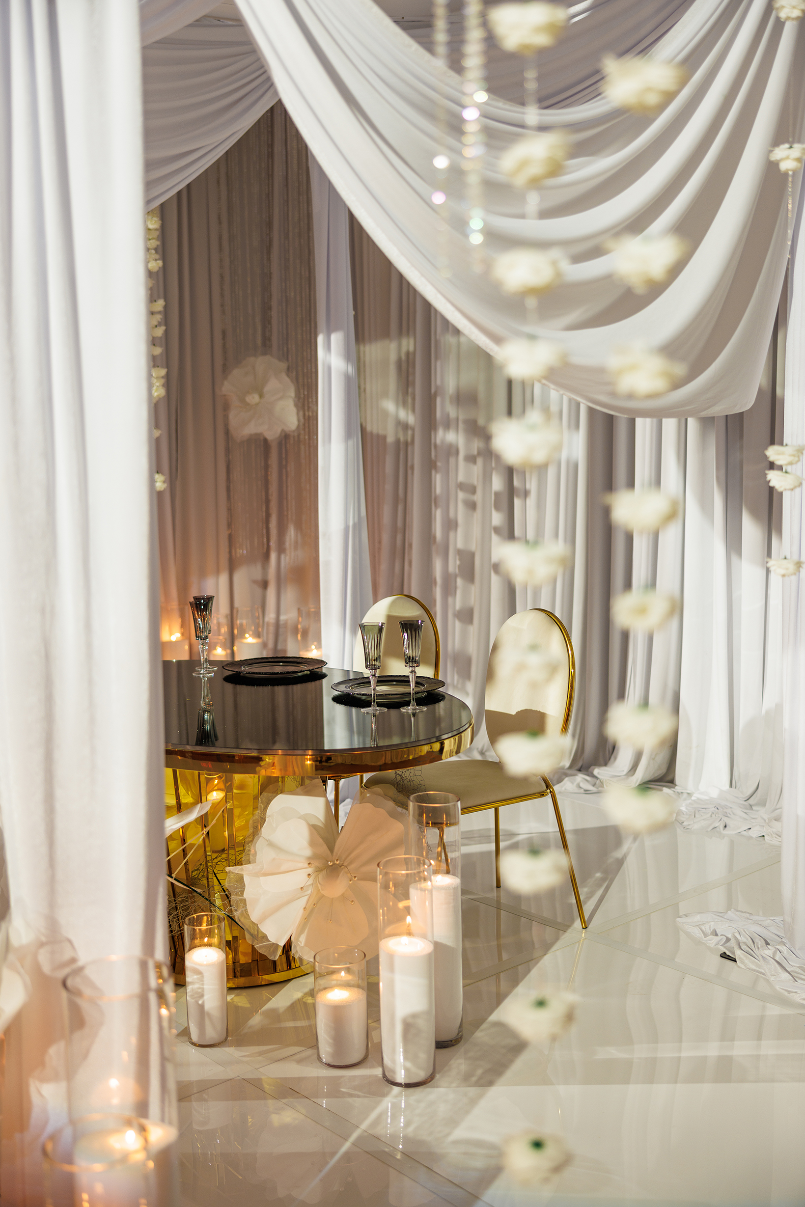 A romantic table for two with gold chairs is set under elegant white drapes, surrounded by candles in glass holders and hanging white flowers, creating a soft, intimate ambiance.