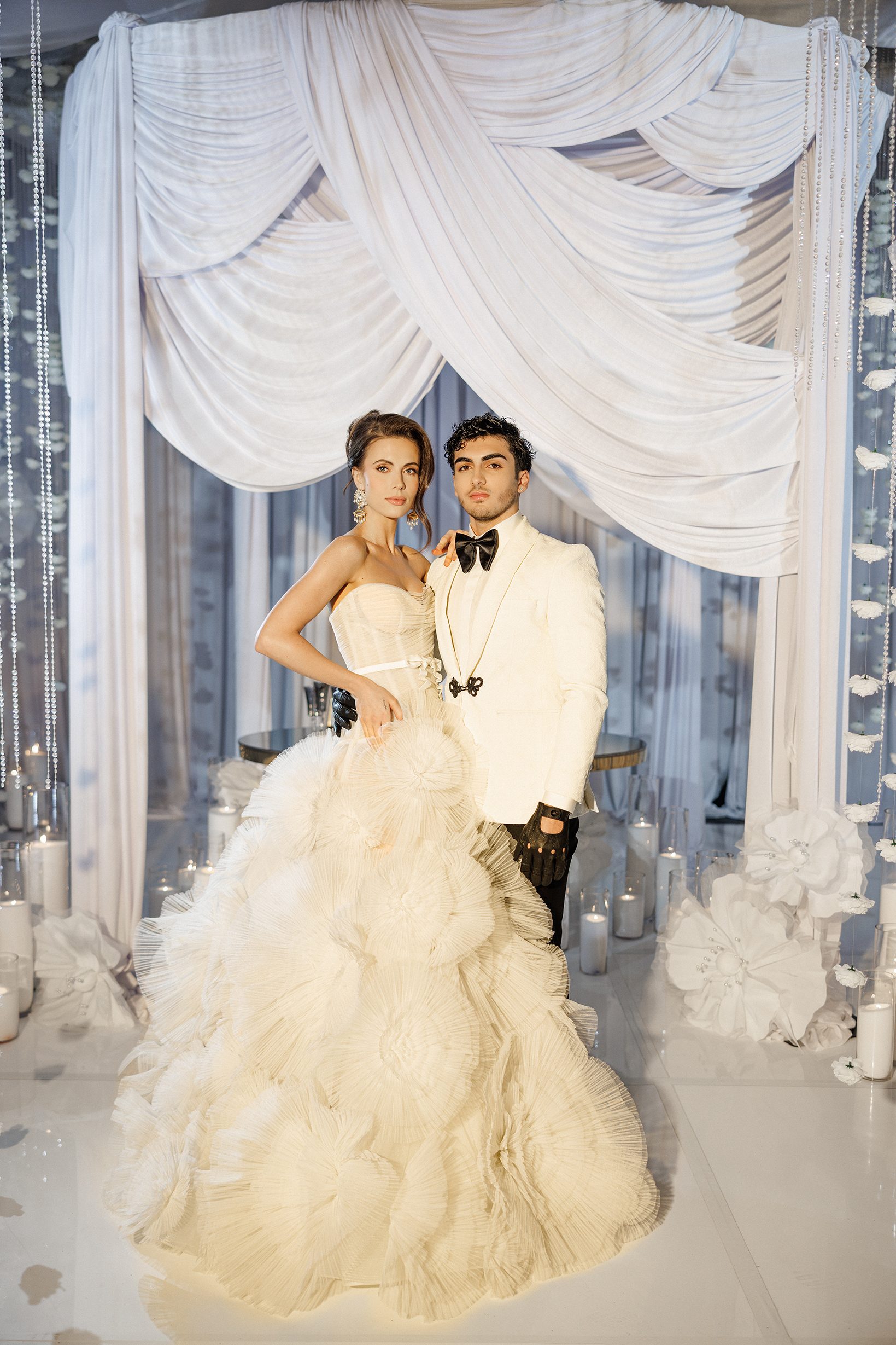 A bride and groom in elegant white attire stand together in front of draped white fabric and candles, creating a glamorous wedding scene. The bride wears a voluminous gown, and the groom wears a tuxedo with black gloves.