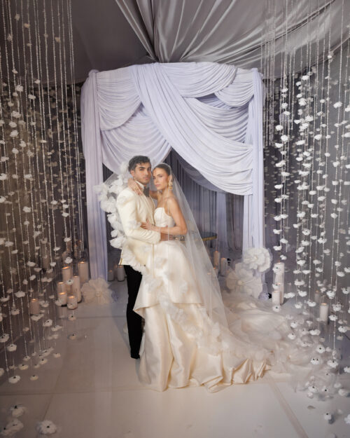 A bride and groom stand together inside a white, flower-filled wedding venue with draped curtains, hanging floral decorations, and candles. The bride wears a long white gown and veil, and the groom wears a cream-colored jacket.