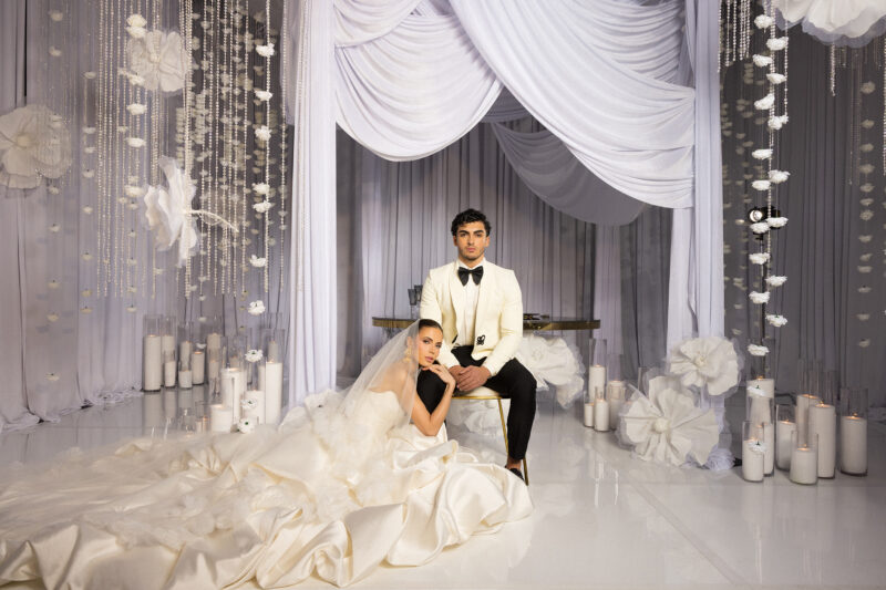 A bride in a voluminous white gown and veil sits on the floor beside a groom in a white tuxedo jacket and black bow tie, surrounded by white drapery, hanging flowers, and lit candles in an elegant wedding setting.