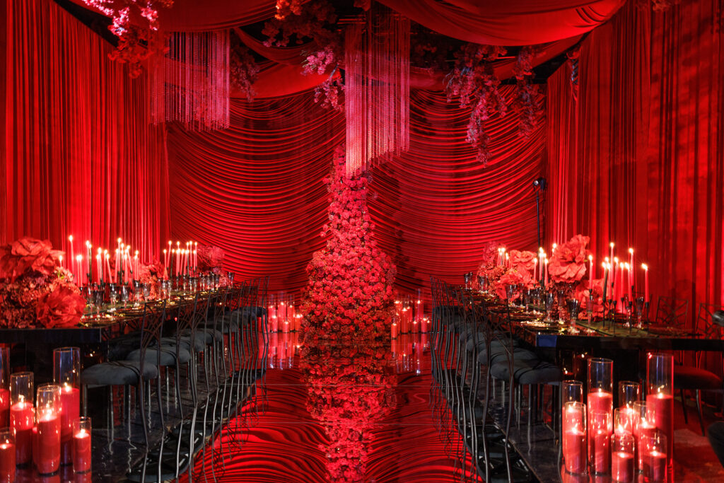 A luxurious event venue draped in red fabric, featuring a dramatic floral centerpiece, long dining tables with black chairs, red candles, and elegant red floral arrangements, creating a romantic and opulent atmosphere.