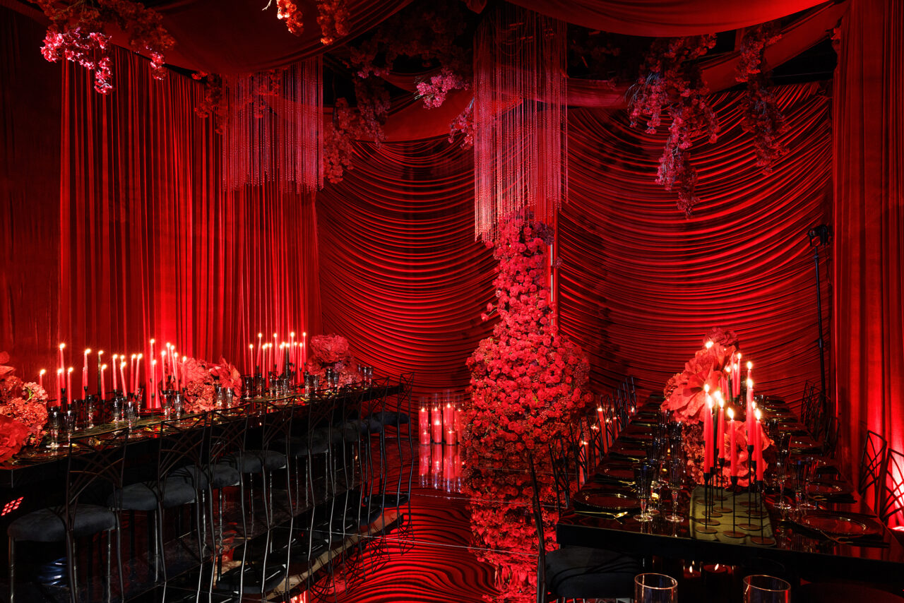 A lavish dining room draped in red fabric and lit by numerous red candles, with floral arrangements and black chairs around elegant, glossy tables, creating a dramatic and luxurious atmosphere.