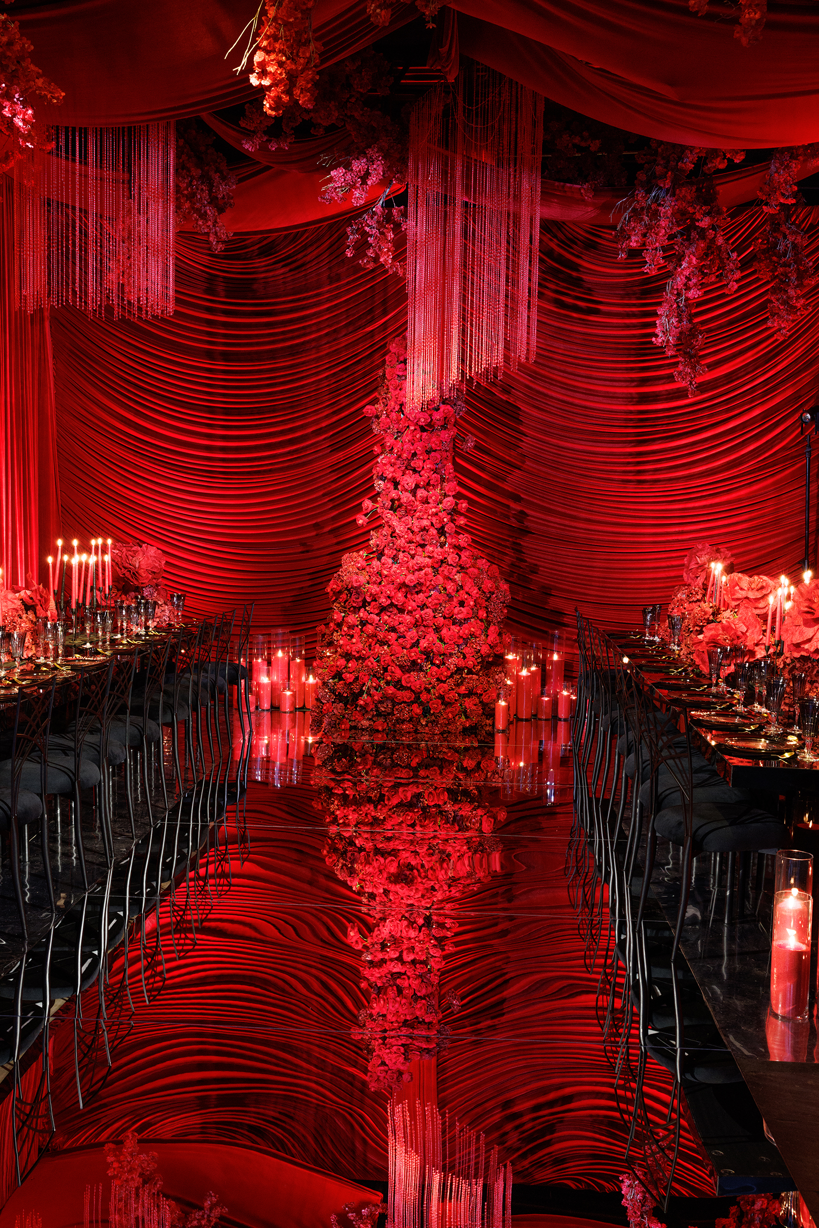 Extravagant dining area draped in red fabric with a central pillar covered in red flowers, candles on long banquet tables, and reflective black flooring creating a dramatic, romantic atmosphere.