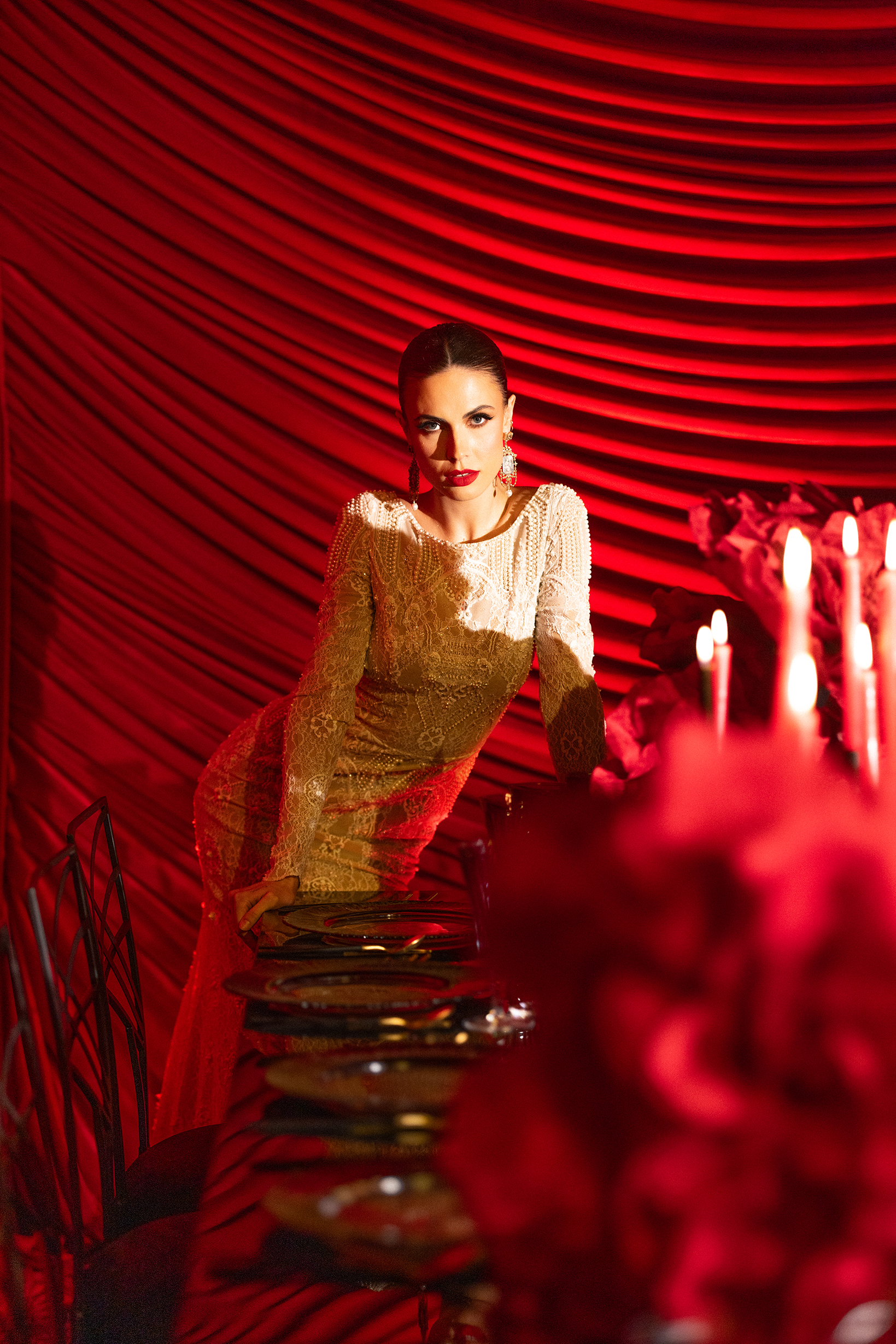 A woman in a gold lace dress poses beside a lavishly set table with red flowers and lit candles, against a backdrop of dramatic red draped fabric. The scene is bathed in warm, red-toned lighting.