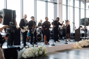 A live band performs onstage at an indoor event, with multiple singers and musicians. White floral arrangements line the stage, and large windows let in natural light.