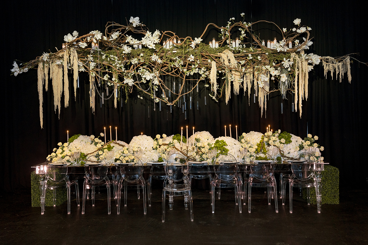 An elegant dining setup with a transparent table and chairs, adorned with large bouquets of white flowers, under a suspended floral arrangement featuring white blooms and hanging crystals.